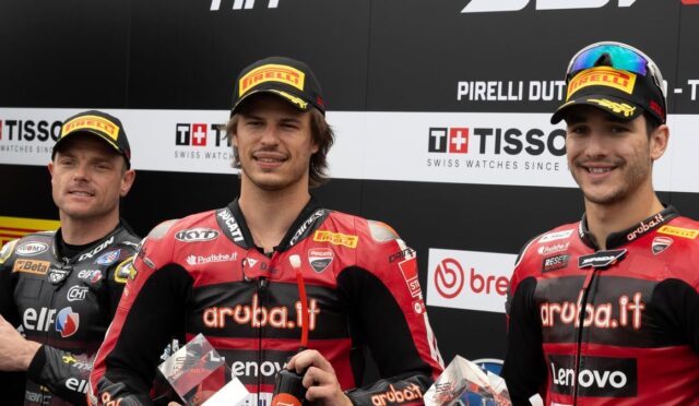 Three motorcycle racers stand on a podium in red-and-black 'aruba.it' racing suits and Pirelli caps, holding trophies against sponsor banners.