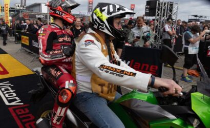 Two motorcycle racers sit on a green Kawasaki at a busy race event, with fans and sponsor banners in the background.