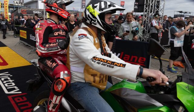 Two motorcycle racers sit on a green Kawasaki at a busy race event, with fans and sponsor banners in the background.