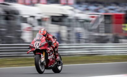 Rider in red racing suit on a red Ducati motorcycle leaning into a turn on a race track, blurred background shows stands and barriers.