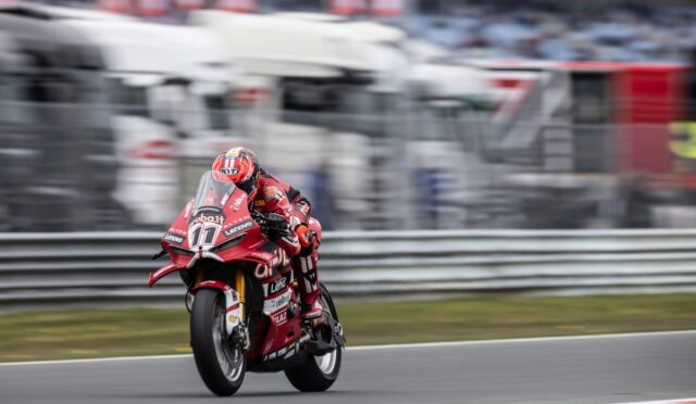 Rider in red racing suit on a red Ducati motorcycle leaning into a turn on a race track, blurred background shows stands and barriers.