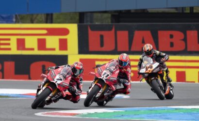 Three motorcycle racers lean into a sharp turn on a race track, red bikes leading the pack with a sponsor-banner wall in the background.