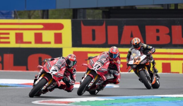 Three motorcycle racers lean into a sharp turn on a race track, red bikes leading the pack with a sponsor-banner wall in the background.