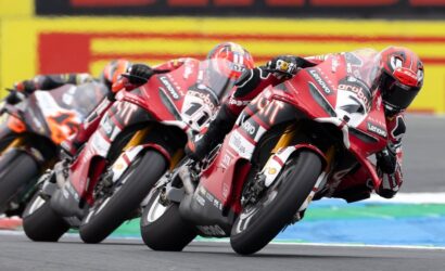 Red motorcycle racers lean into a turn in a high-speed MotoGP-style race on a track nearby the camera edge.