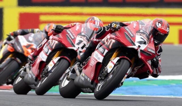 Red motorcycle racers lean into a turn in a high-speed MotoGP-style race on a track nearby the camera edge.