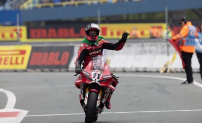 Moto racer on a red bike crosses the line, raises a gloved fist in victory on a grey track with Pirelli boards in the background.