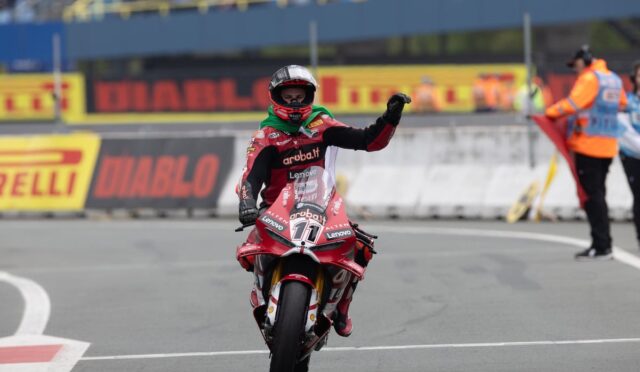 Moto racer on a red bike crosses the line, raises a gloved fist in victory on a grey track with Pirelli boards in the background.