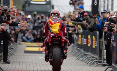 Motocycle racer in red suit rides away from camera on a crowd-lined finish area, spectators reach out and cheer with phones upclouded by barriers.