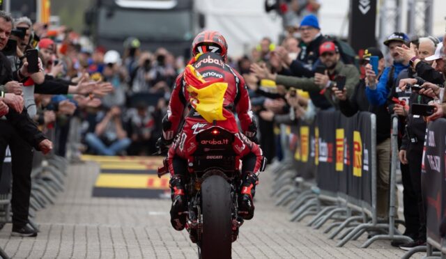 Motocycle racer in red suit rides away from camera on a crowd-lined finish area, spectators reach out and cheer with phones upclouded by barriers.