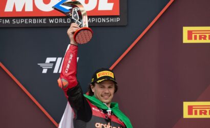 Racer on the podium raises a trophy, wearing a Pirelli cap and Aruba.it racing suit with a green scarf