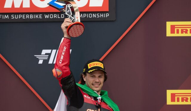 Racer on the podium raises a trophy, wearing a Pirelli cap and Aruba.it racing suit with a green scarf