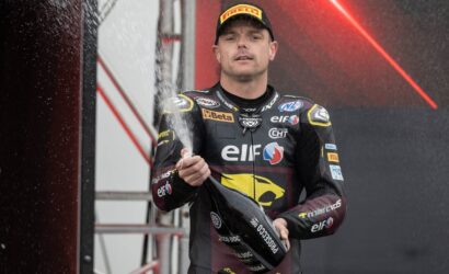 Racing driver in a black and yellow ELF suit sprays champagne on a podium celebrate win, holding a bottle with a focused expression