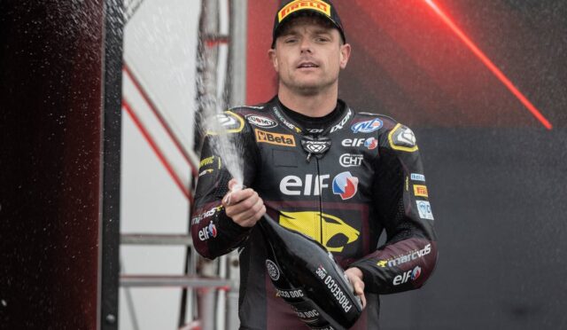 Racing driver in a black and yellow ELF suit sprays champagne on a podium celebrate win, holding a bottle with a focused expression