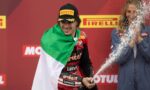 Motorsport rider on a podium, wrapped in an Italian flag, spraying champagne with a smile against a Pirelli backdrop