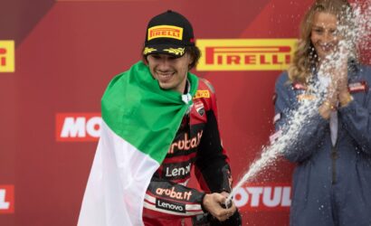 Motorsport rider on a podium, wrapped in an Italian flag, spraying champagne with a smile against a Pirelli backdrop