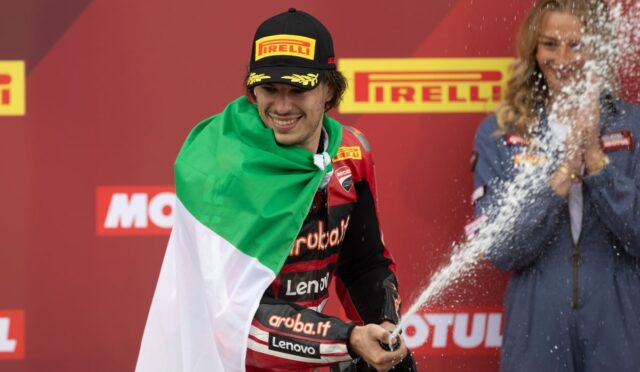 Motorsport rider on a podium, wrapped in an Italian flag, spraying champagne with a smile against a Pirelli backdrop