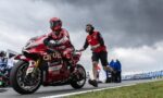Moto rider in red racing suit on a red sport bike, being guided on a race track by a crew member in a red shirt against a cloudy sky.