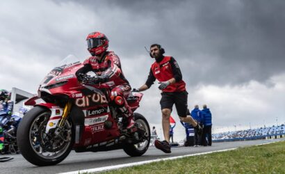 Moto rider in red racing suit on a red sport bike, being guided on a race track by a crew member in a red shirt against a cloudy sky.