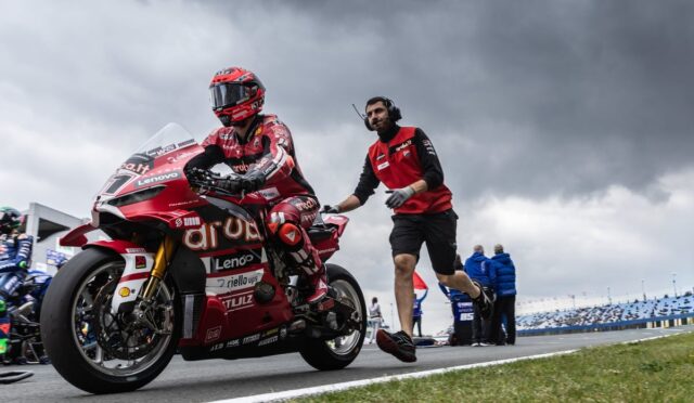 Moto rider in red racing suit on a red sport bike, being guided on a race track by a crew member in a red shirt against a cloudy sky.