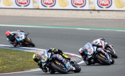 Motorcycle racers lean into a sharp turn on a race track; foreground rider #55 in blue with neon yellow gloves leads the group.