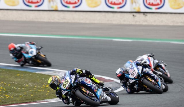 Motorcycle racers lean into a sharp turn on a race track; foreground rider #55 in blue with neon yellow gloves leads the group.