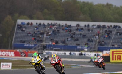 Two motorcycle racers lean into a curve on a race track while crowds watch from grandstands in the background, bike #34 in yellow and white leading #5 in red and black.
