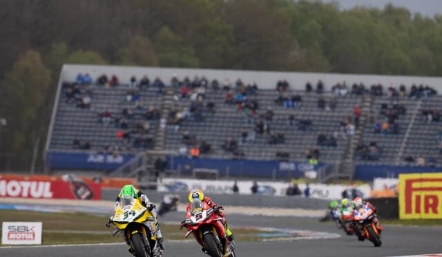 Two motorcycle racers lean into a curve on a race track while crowds watch from grandstands in the background, bike #34 in yellow and white leading #5 in red and black.
