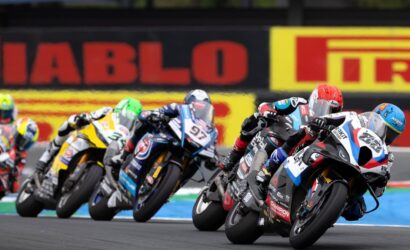 Group of motorcycle racers leaning into a curve on a race track, colorful bikes and helmets in a tight pack under trackside banners