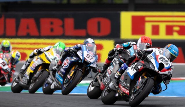 Group of motorcycle racers leaning into a curve on a race track, colorful bikes and helmets in a tight pack under trackside banners