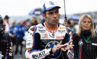 MotoGP rider in racing suit with BMW logos, blue cap, clapping hands in a crowded paddock.