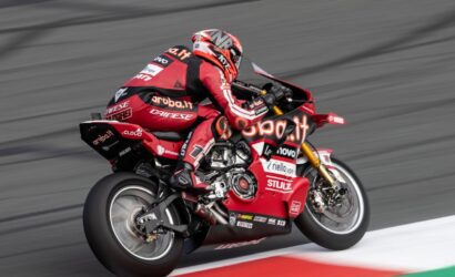 MotoGP rider on a red Ducati racing bike leaning into a turn on a race track, sponsor logos visible across suit and bike