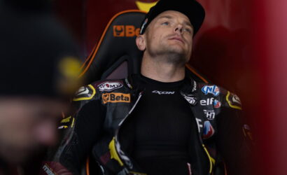Racer in a black racing suit with sponsor patches sits in an orange-and-black seat, looking upward in the pit area.