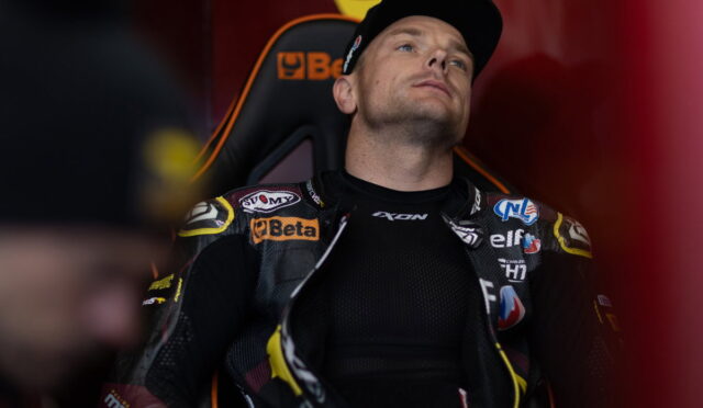 Racer in a black racing suit with sponsor patches sits in an orange-and-black seat, looking upward in the pit area.