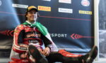 Motorsport racer in red racing suit sits on the floor with a green and white scarf, smiling in front of sponsor backdrop.