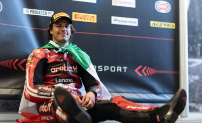 Motorsport racer in red racing suit sits on the floor with a green and white scarf, smiling in front of sponsor backdrop.
