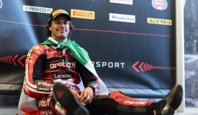 Motorsport racer in red racing suit sits on the floor with a green and white scarf, smiling in front of sponsor backdrop.