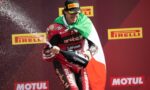 Racer on a podium sprays champagne while draped in the Italian flag, with Pirelli and Motul branding in the background.