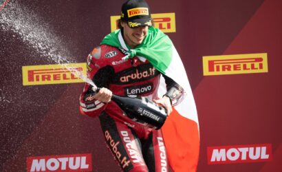 Racer on a podium sprays champagne while draped in the Italian flag, with Pirelli and Motul branding in the background.