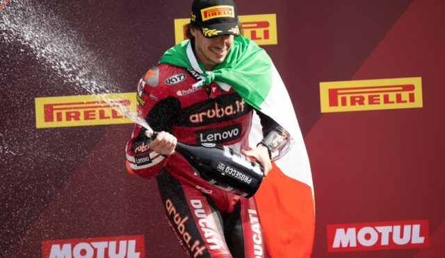 Racer on a podium sprays champagne while draped in the Italian flag, with Pirelli and Motul branding in the background.