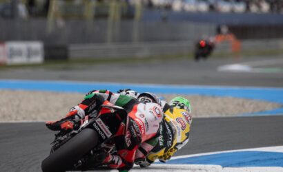 Two motorcycle racers lean into a sharp turn on a race track, close behind each other in colorful racing suits and bikes.