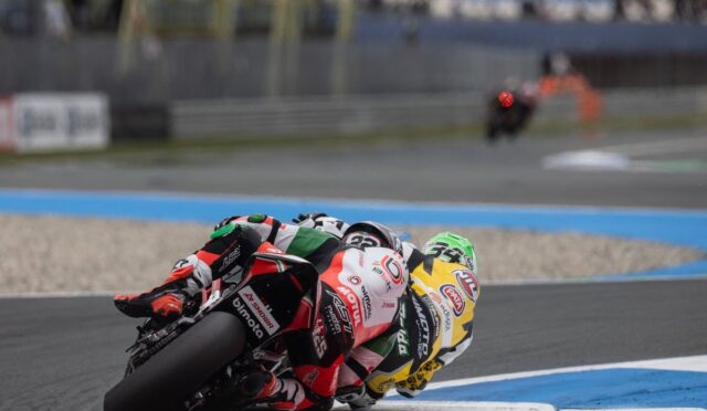 Two motorcycle racers lean into a sharp turn on a race track, close behind each other in colorful racing suits and bikes.