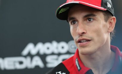 Close-up of a young racing driver in a red cap and team polo, speaking during a media interview at a race event.