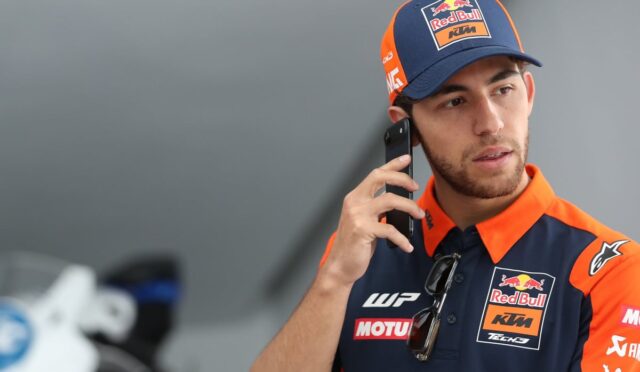 Rider in Red Bull KTM team uniform talking on a cellphone outdoors or in a paddock area.