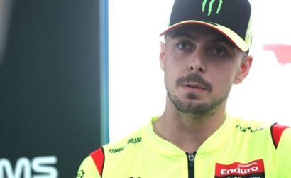 Portrait of a male motorcycle racer in a neon-yellow racing jacket and black cap with a green Monster logo, looking to the side.