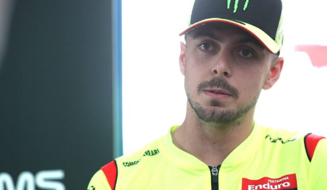 Portrait of a male motorcycle racer in a neon-yellow racing jacket and black cap with a green Monster logo, looking to the side.