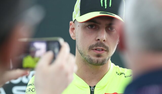 Man in neon yellow racing gear being photographed; focused expression, cap with green Monster logo in a media interview scene