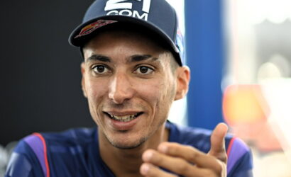 Close-up of a man wearing a dark cap and blue athletic shirt, smiling and gesturing with his hand toward the camera.