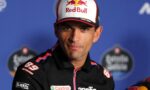 Racing driver in a Red Bull cap and black Aprilia jacket at a press conference with sponsor logos visible.