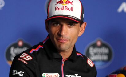 Racing driver in a Red Bull cap and black Aprilia jacket at a press conference with sponsor logos visible.