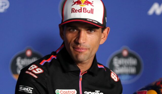 Racing driver in a Red Bull cap and black Aprilia jacket at a press conference with sponsor logos visible.